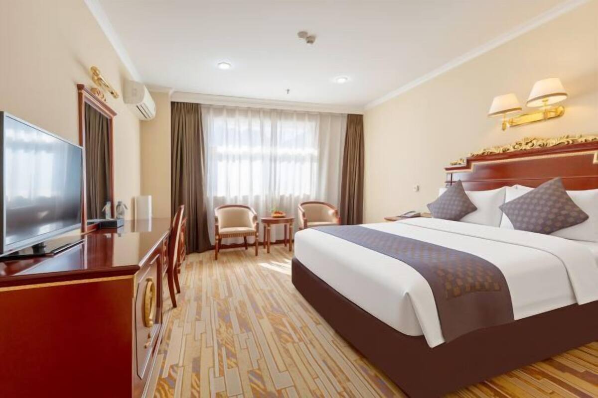 A hotel room with a large bed, a desk with a television, two chairs, a small table, and a window with curtains.