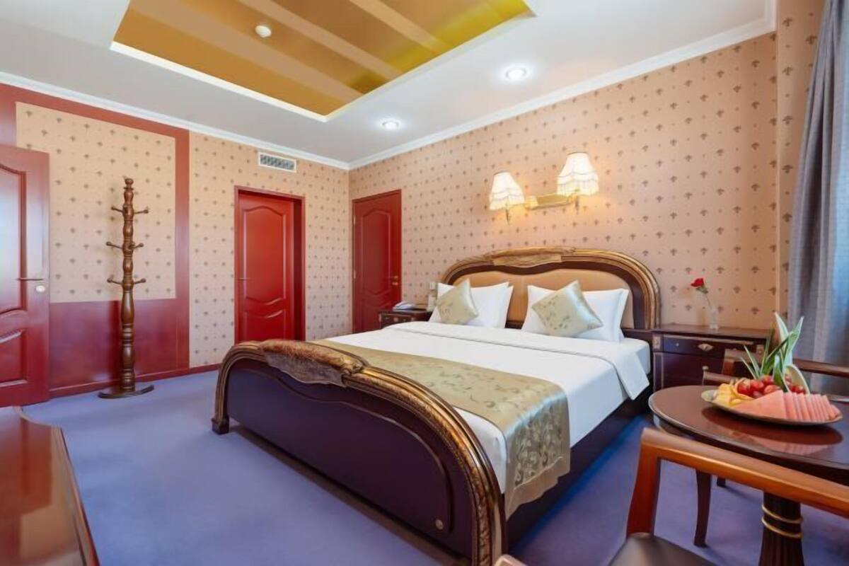 A hotel room with a bed, a desk, a chair, a coat rack, and a wall-mounted lamp.