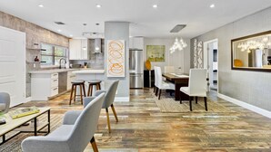 A modern kitchen with white cabinets, a stainless steel refrigerator, and a dining area with a large mirror and chandelier. - La Bougainvillea: 3BR, 2BA Home - Heated Pool (Fort Lauderdale)