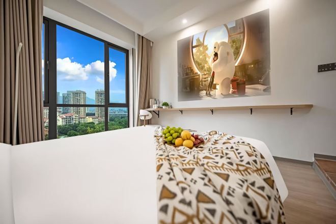 A hotel room with a large window, a bed with a patterned bedspread, a fruit bowl on the bed, a wall-mounted shelf with a decorative item, and a view of buildings outside.