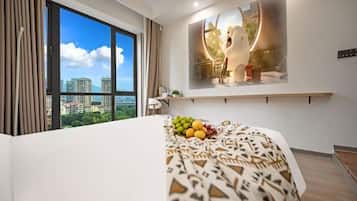 A hotel room with a large window, a bed with a patterned bedspread, a fruit bowl on the bed, a wall-mounted shelf with a decorative item, and a view of buildings outside.