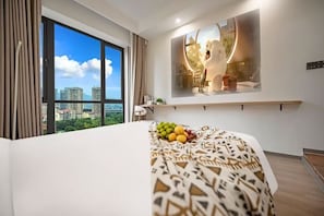 A hotel room with a large window, a bed with a patterned bedspread, a fruit bowl on the bed, a wall-mounted shelf with a decorative item, and a view of buildings outside. - Yiting Hotel (Chongqing)