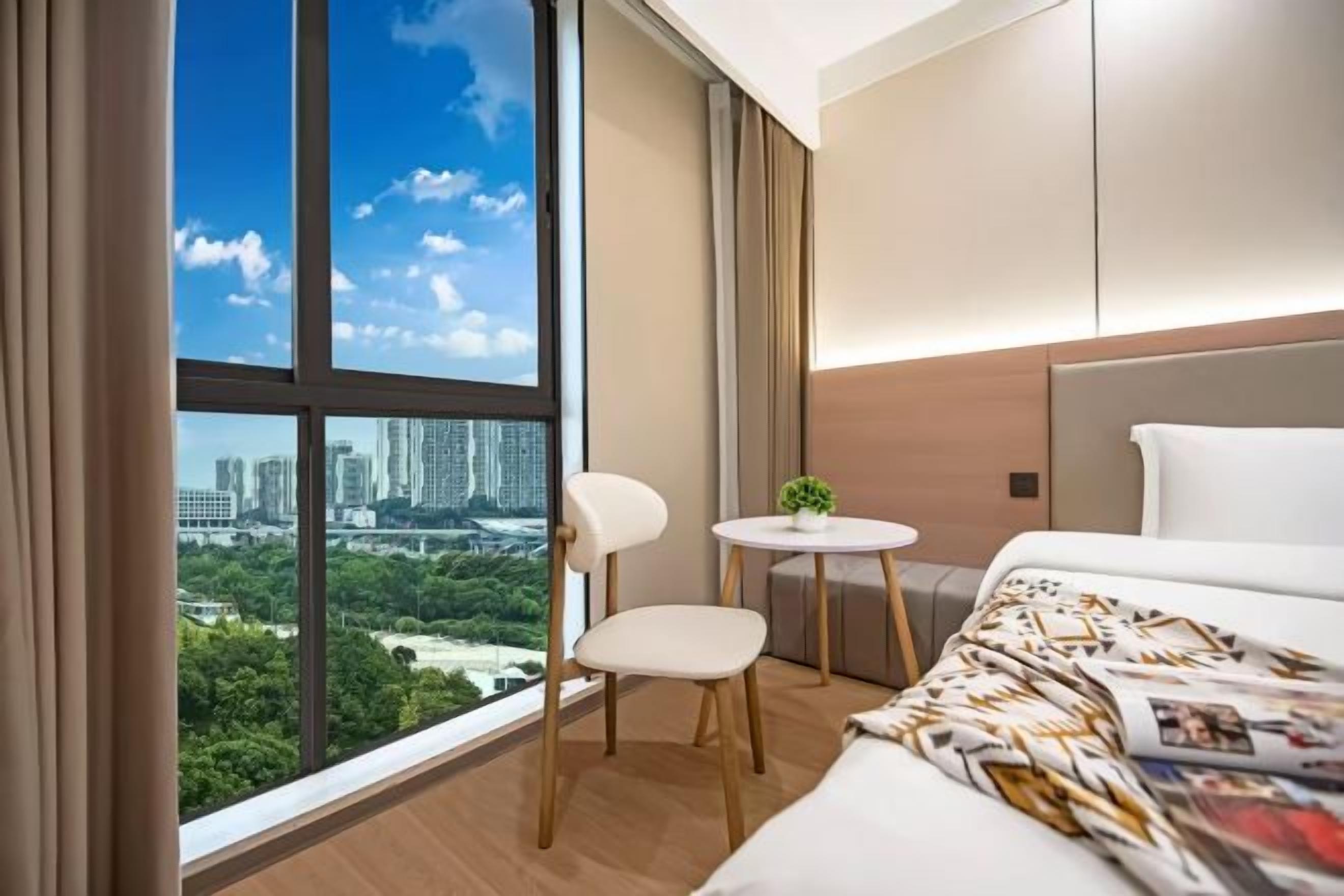 A hotel room with a large window, a bed, a chair, a small table with a plant, and a view of a cityscape.