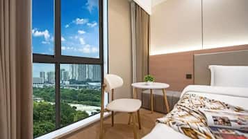 A hotel room with a large window, a bed, a chair, a small table with a plant, and a view of a cityscape.