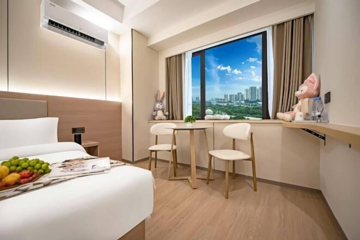 Habitación de hotel con cama, un pequeño comedor con sillas, ventana con vista a la ciudad y un equipo de aire acondicionado.