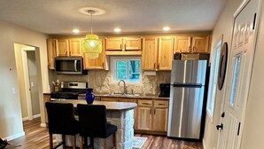 Cocina compacta con armarios de madera, microondas integrado, refrigerador de acero inoxidable y comedor con una silla de mimbre y dos sillas.