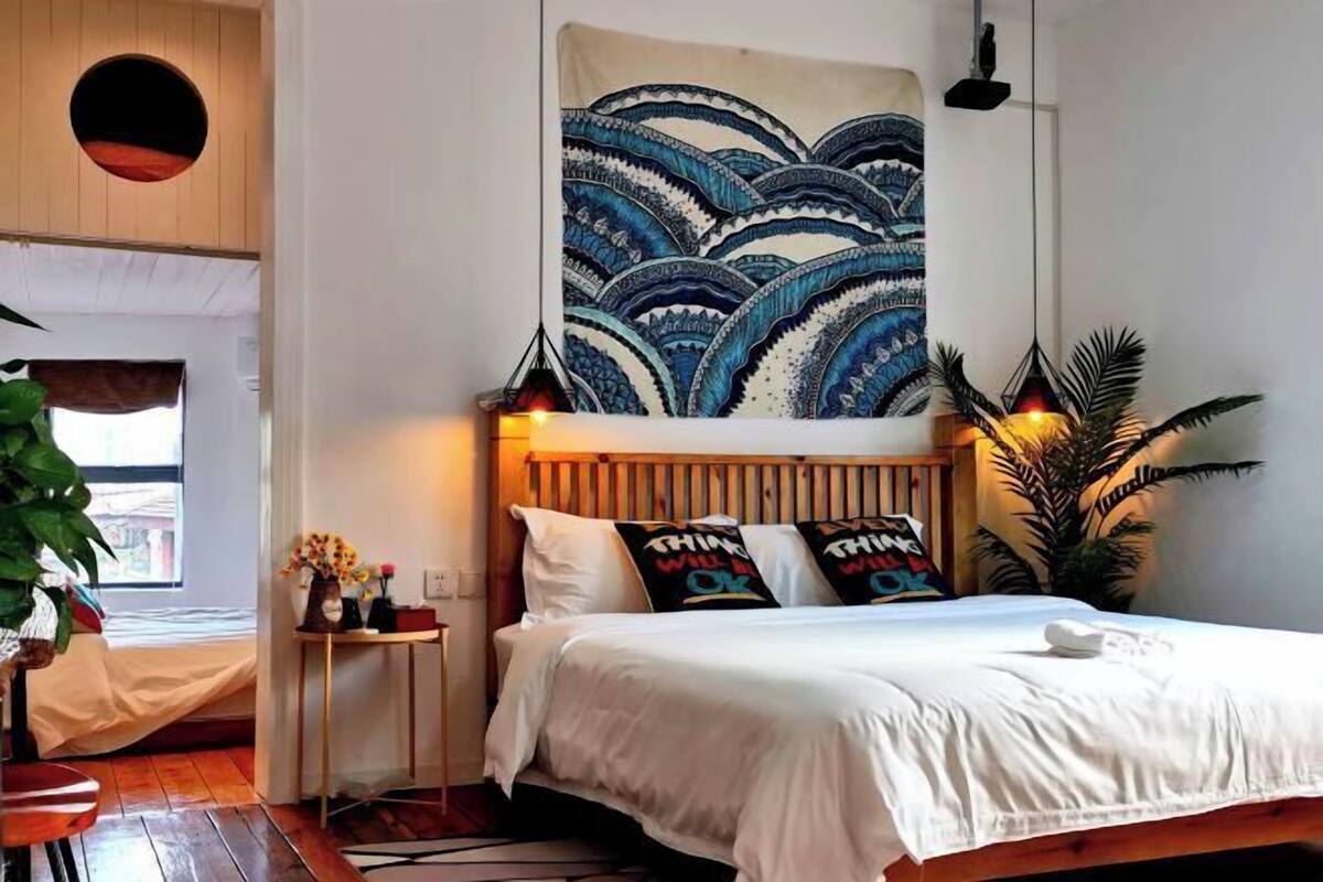 A bedroom with a large bed, a wooden headboard, a wall tapestry, and a small bedside table with a lamp and flowers.