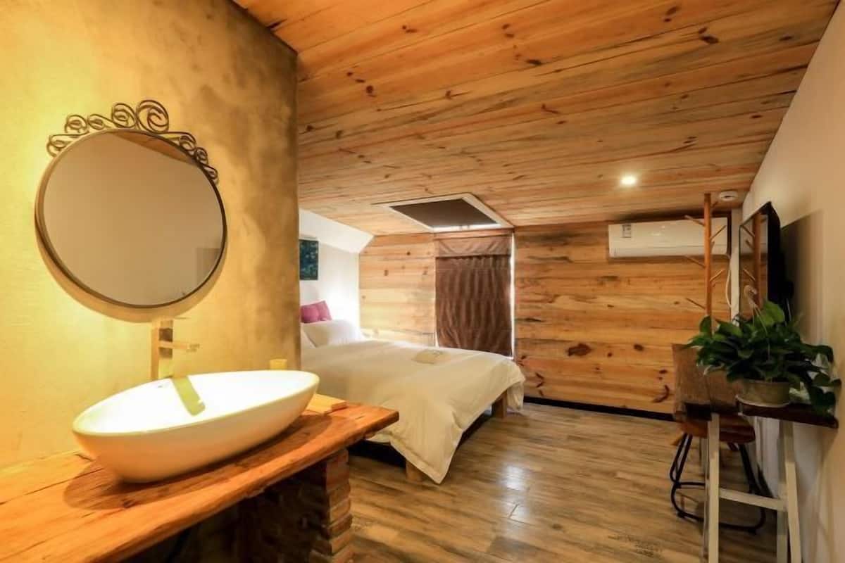 A wooden-paneled room with a large round mirror, a white sink, a bed, and a small table with a plant.