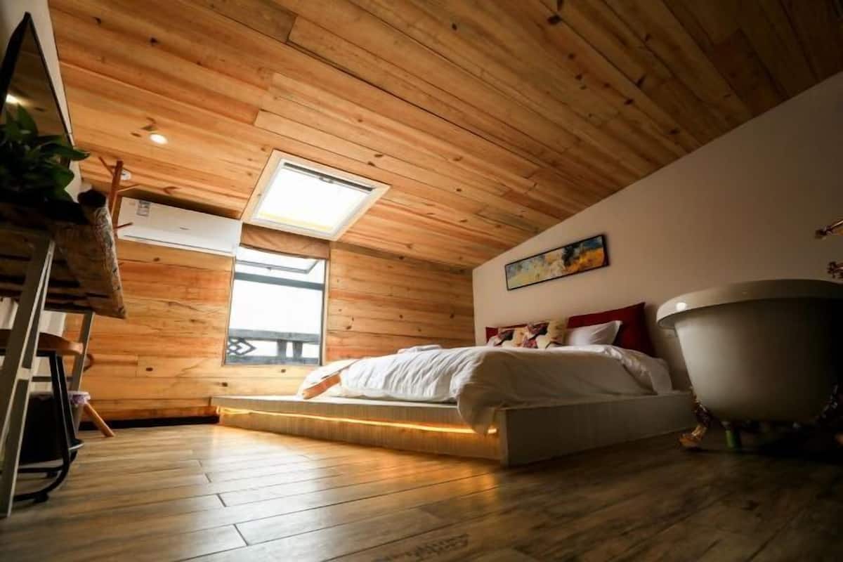 A bedroom with a wooden ceiling, a skylight, a bed, a bathtub, and a window.