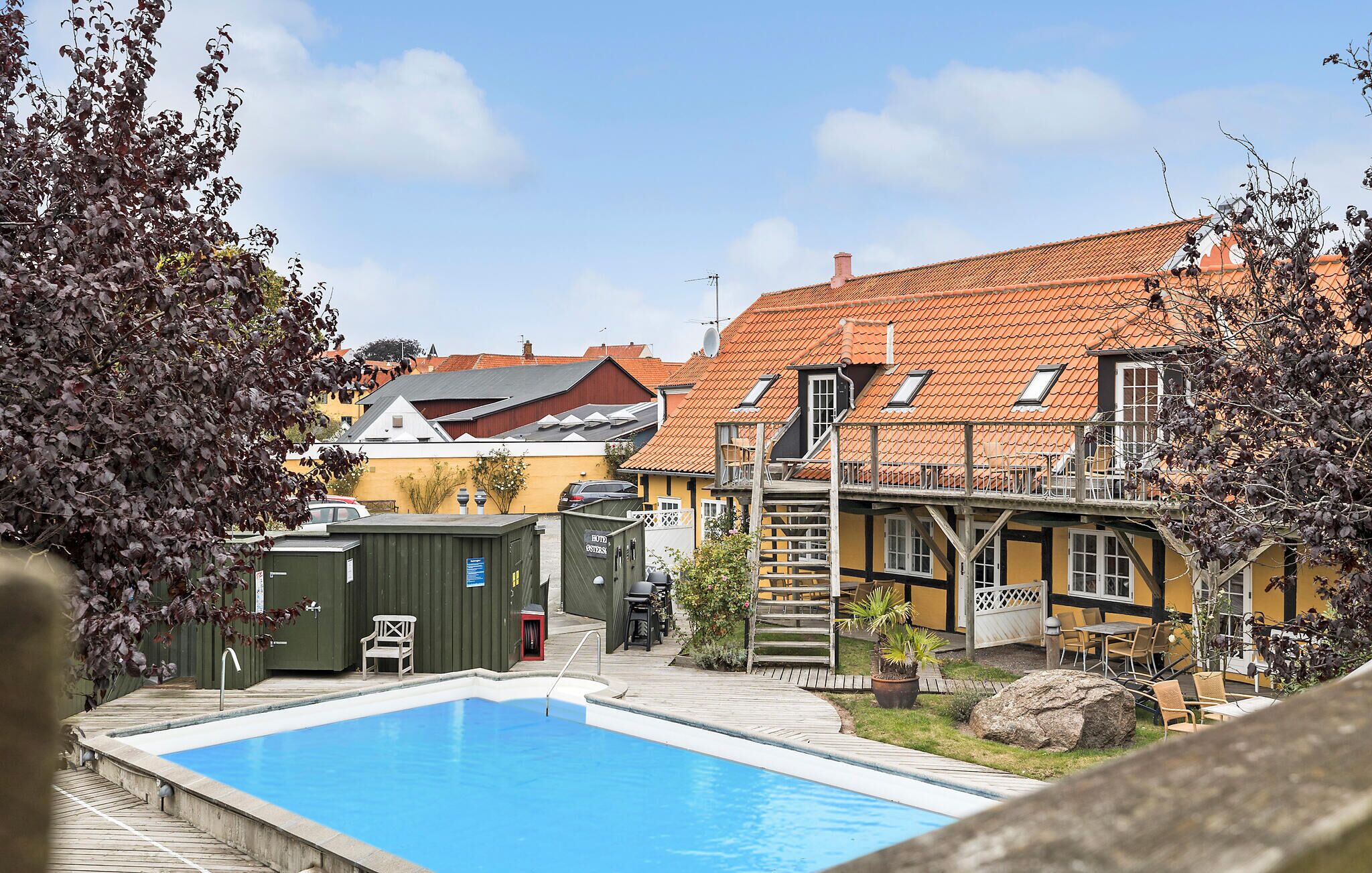 Un espace piscine avec une terrasse en bois, un petit jardin et un bâtiment doté d’un toit recouvert de tuiles rouges.