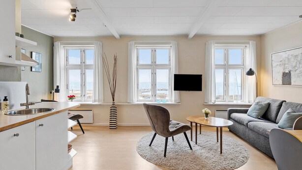 En moderne stue med en grå sofa, et rundt sofabord og en grå lænestol. To vinduer med udsigt til havet og et fjernsyn monteret på væggen.