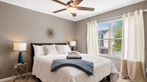 Un dormitorio con cama, ventilador de techo, lámparas de noche y una ventana con cortinas.