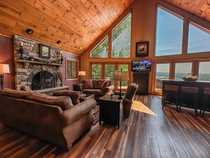 A cozy living room with a stone fireplace, leather sofas, a TV, and large windows offering a scenic view.
