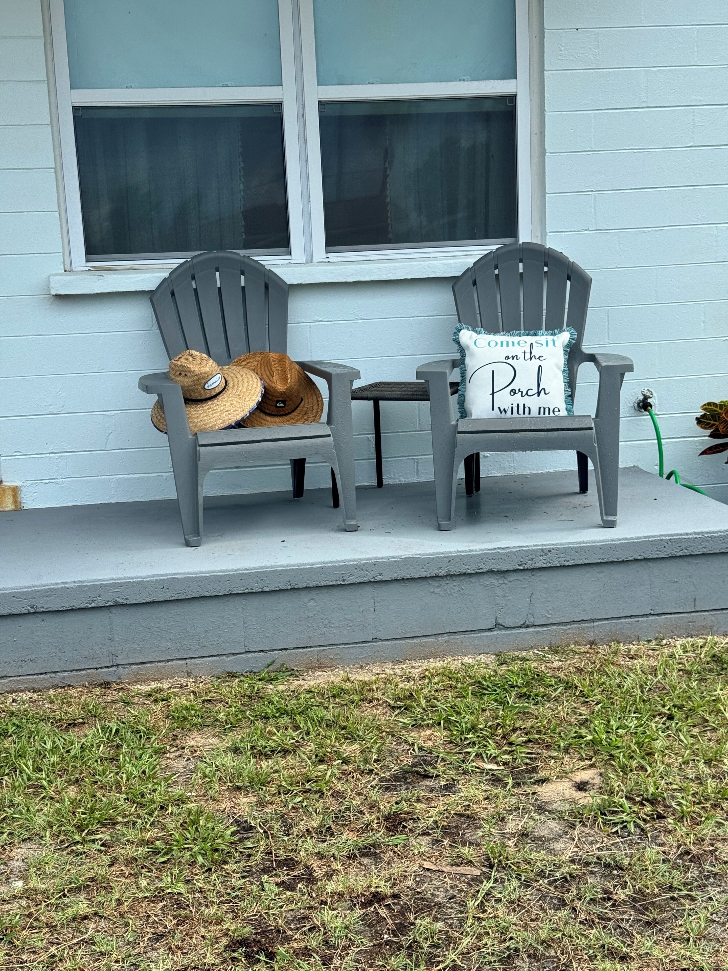Zwei graue Adirondack-Stühle auf einer Veranda mit einer weißen Wand und einem Fenster im Hintergrund.