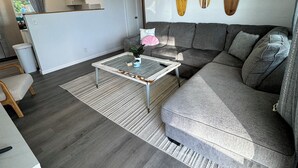 A modern living room with a gray sofa, a coffee table, and a dining area with a white table and chairs. There are surfboards on the wall, a ceiling fan, and a window with curtains. - It's beach time! Enjoy the sunshine! (Ormond Beach)