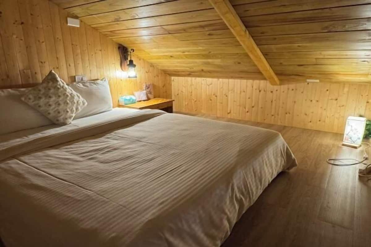 A cozy attic bedroom with a wooden ceiling, a large bed with a striped bedspread, a nightstand, and a wall-mounted lamp.