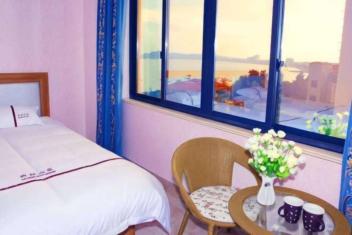 Een hotelkamer met een bed, een rieten stoel, een klein tafeltje met een bloevas en uitzicht op de stad door grote ramen.