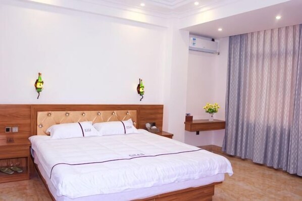 A hotel room with a large bed, bedside tables, a nightstand, an air conditioning unit, and a window with curtains.