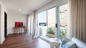Una sala de estar moderna con piso de madera, un sofá blanco, una mesa de comedor con sillas y un gran ventanal corredizo que da a un balcón con vista a un edificio y una masa de agua.