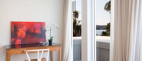 A room with a wooden desk, a white chair, a red abstract painting, a white orchid, and a view of a body of water through a glass door.