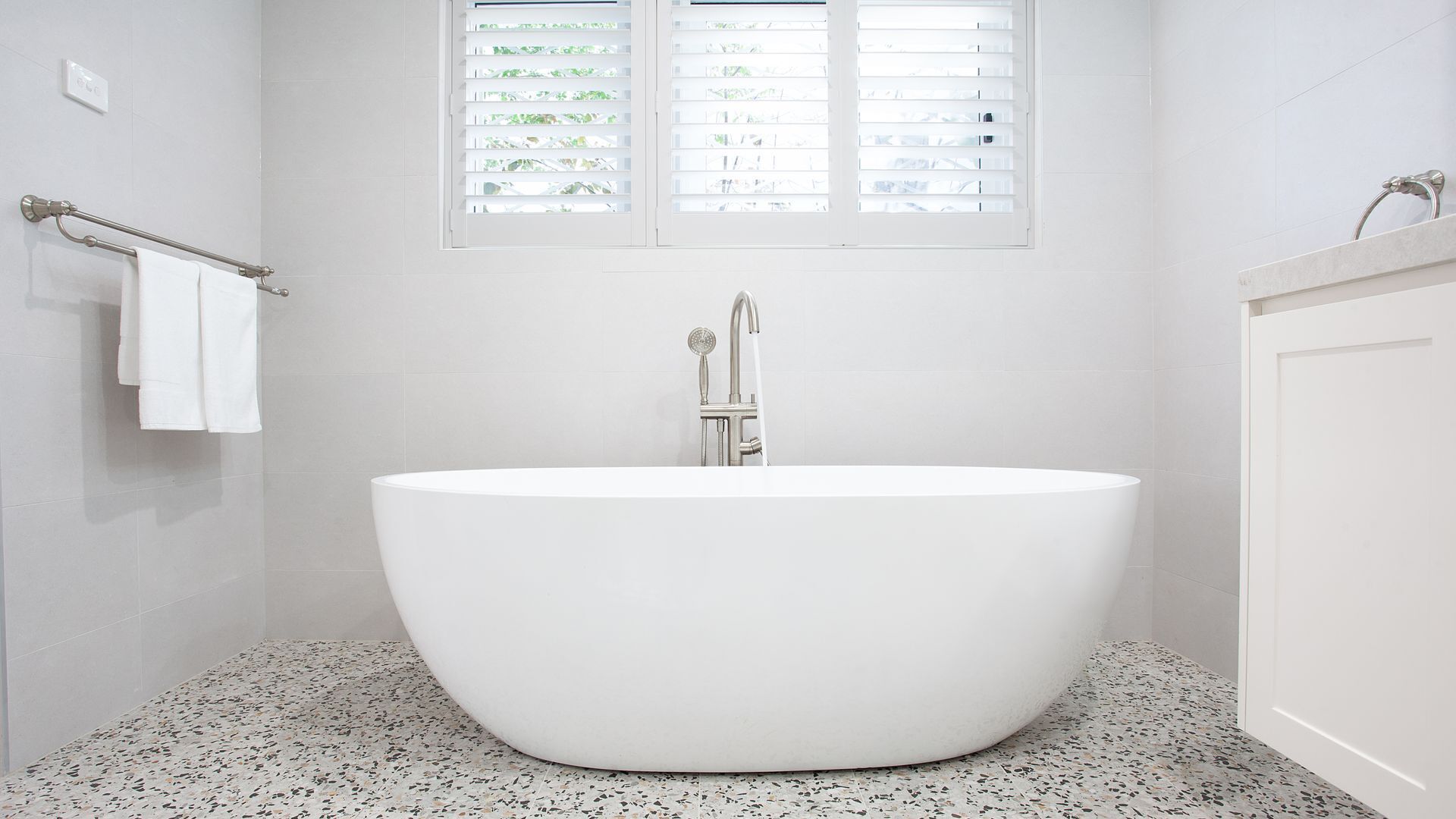Un baño moderno con una bañera independiente, una ventana con persianas y un perchero con toallas blancas.
