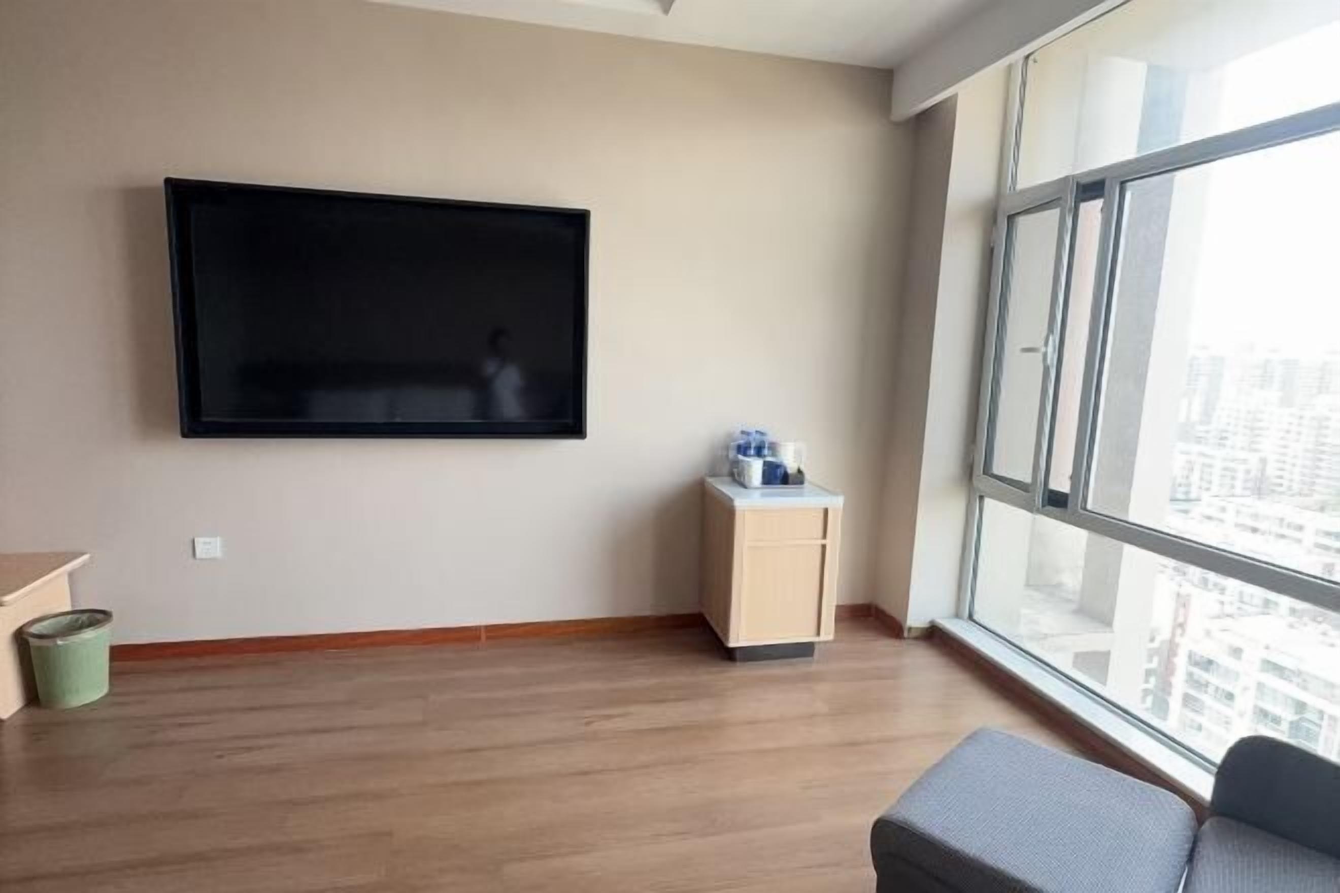 A modern living room with a large flat-screen TV mounted on the wall, a small wooden cabinet, and a large window offering a city view.