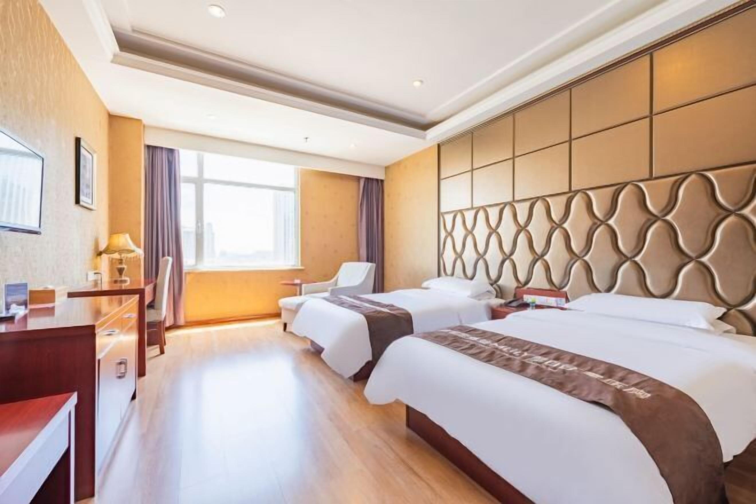 A hotel room with two beds, a desk, a chair, and a large window.