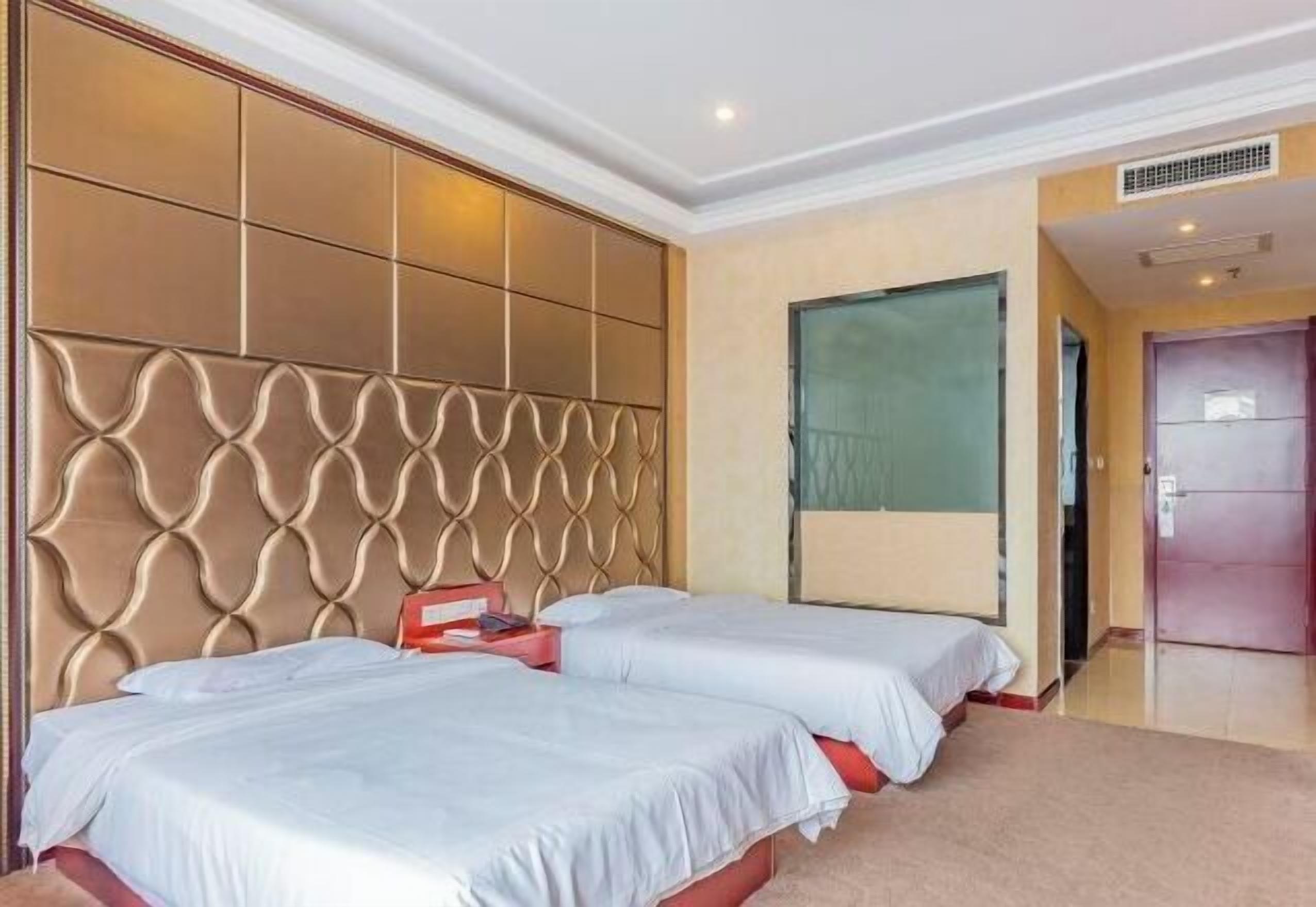 A hotel room with two beds, a large padded headboard, a large window, and a door.