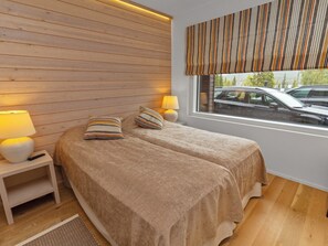 A bedroom with two beds, a nightstand with a lamp, and a window with a view of parked cars. - Holy lake chalet a by Interhome (Kuusamo)