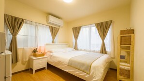 Un dormitorio con cama, mesita de noche, lámpara, equipo de aire acondicionado, cortinas y un pequeño estante de madera.