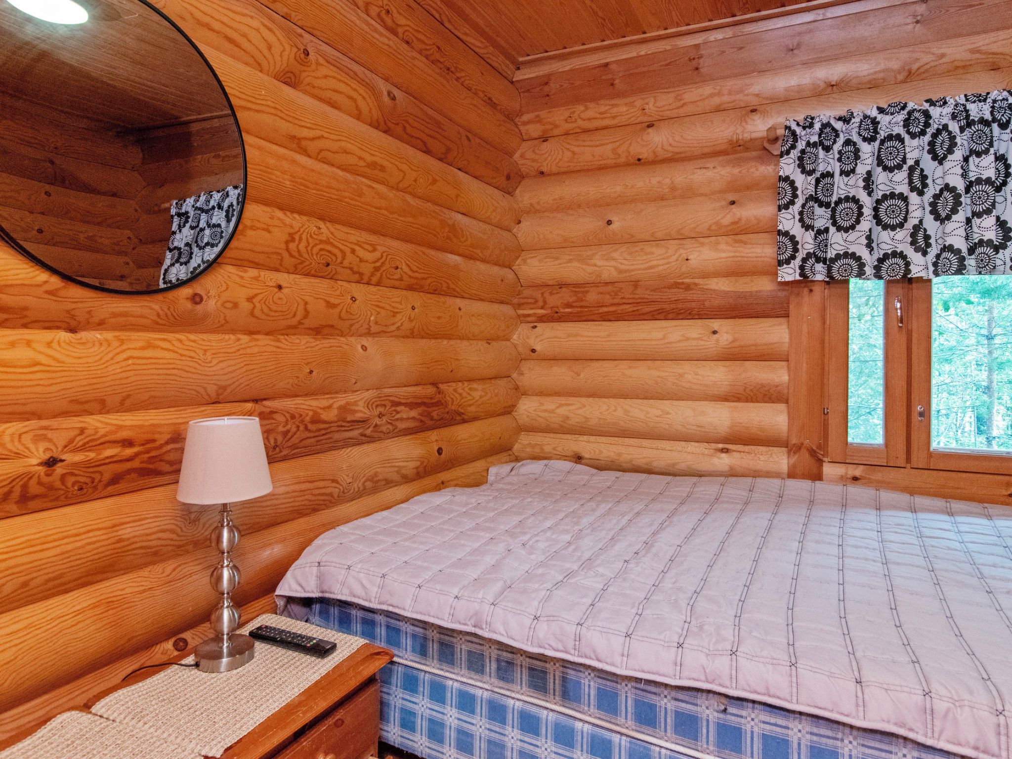 Een houten kamer met een bed, een bedlamp, een spiegel en een raam met gordijnen met een patroon.