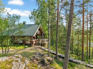 Un chalet rustique avec un toit en métal, entouré d’une forêt et doté d’une terrasse en bois menant à l’entrée.