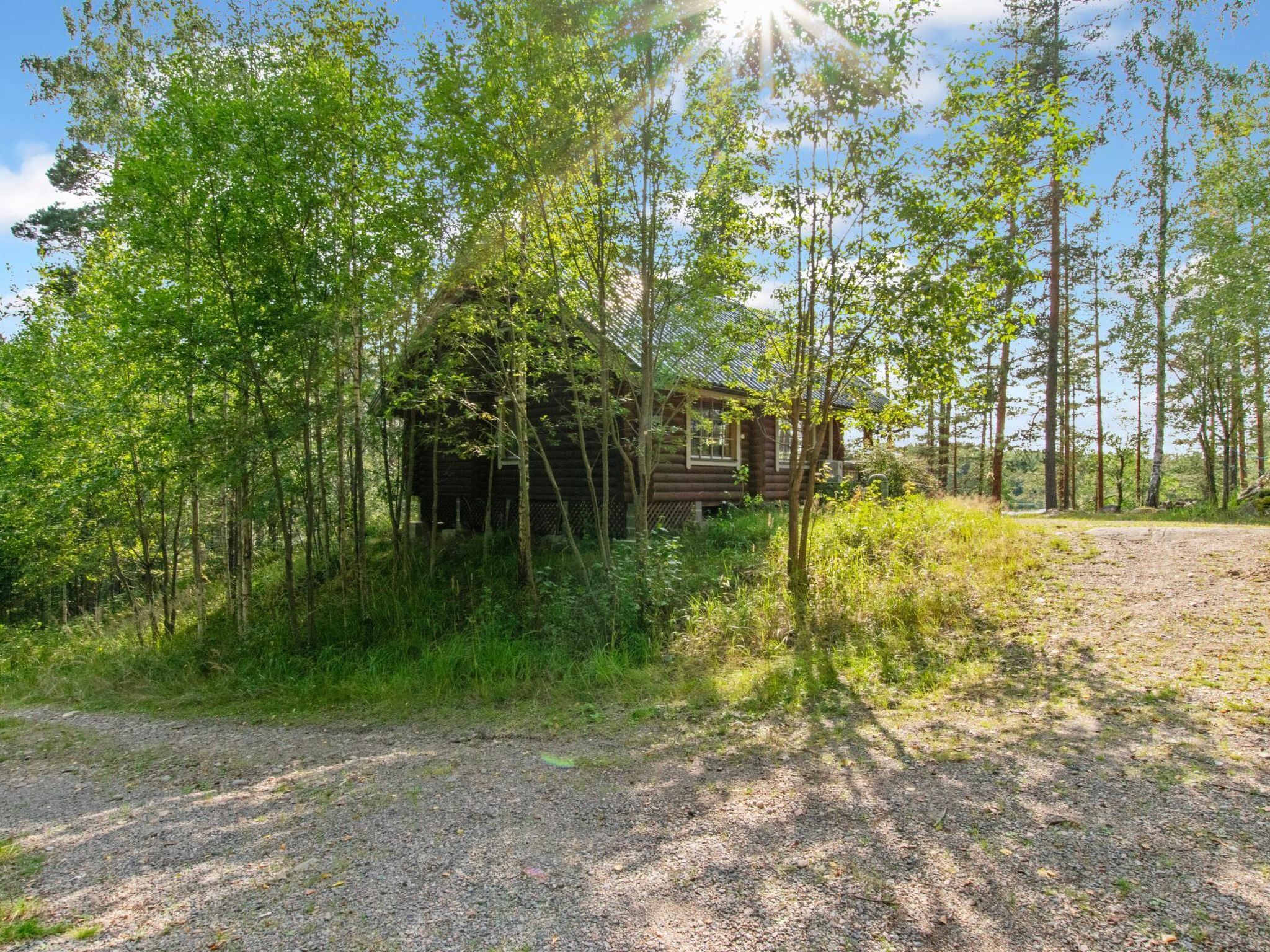Mökki, joka on rakennettu maalaismaisessa tyylissä ja sijaitsee tiheän metsän ympäröimänä. Mökille johtaa sorapolkua.