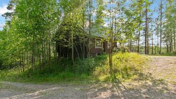 Mökki, joka on rakennettu maalaismaisessa tyylissä ja sijaitsee tiheän metsän ympäröimänä. Mökille johtaa sorapolkua.