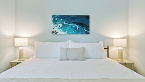 Ein ordentlich bezogenes Bett mit weißen Leinentüchern, zwei Nachttische mit Lampen und ein an der Wand hängendes Bild von einer blauen Meereslandschaft.