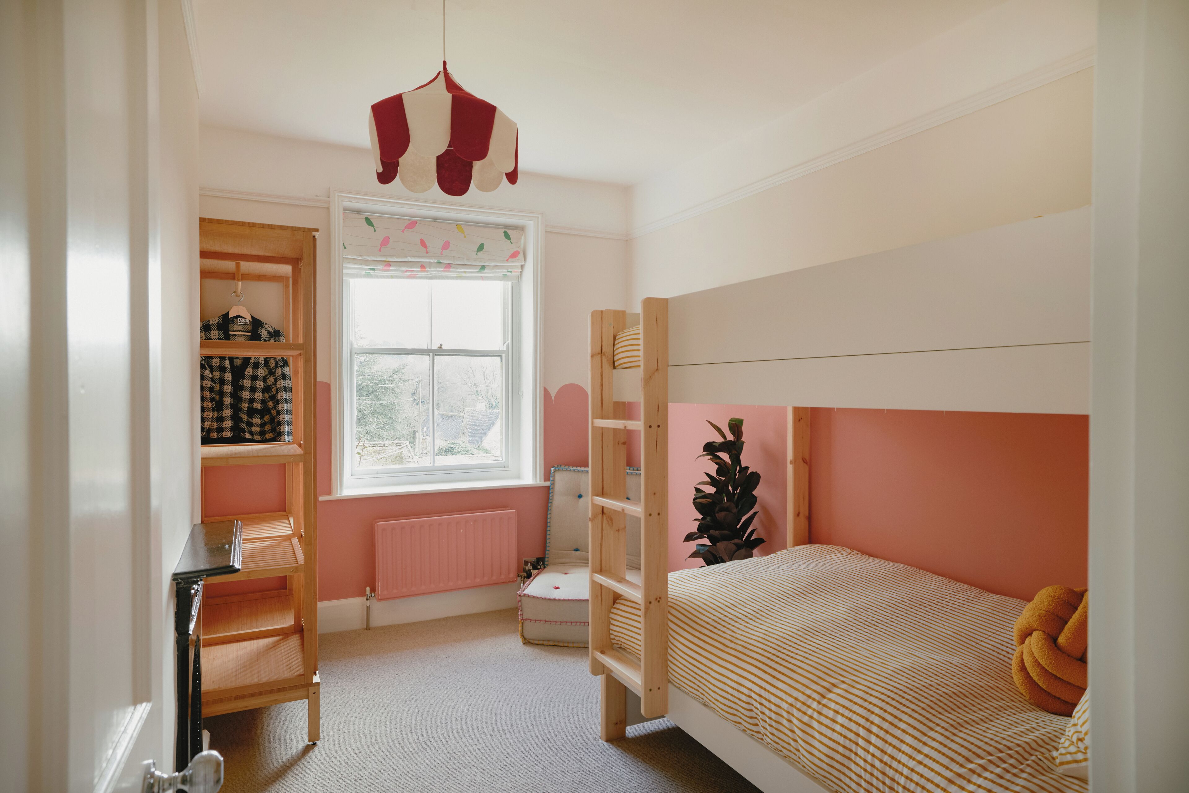 A bedroom with a bunk bed, a desk with shelves, a radiator, and a window with blinds.