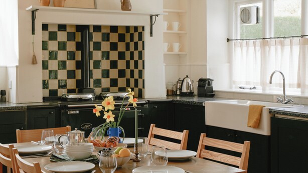 A kitchen with a checkered backsplash, a dining table set with plates and glasses, and a window with a curtain. - Trinity Farmhouse-Stunning House&Garden (Cirencester)