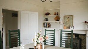A dining area with a white table, green chairs, a vase with flowers, and a bookshelf with books and decorative items. - Trinity Farmhouse-Stunning House&Garden (Cirencester)
