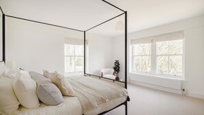 A four-poster bed with a white bedspread and multiple pillows, a white chair, a potted plant, and large windows with white curtains. - Trinity Farmhouse-Stunning House&Garden (Cirencester)