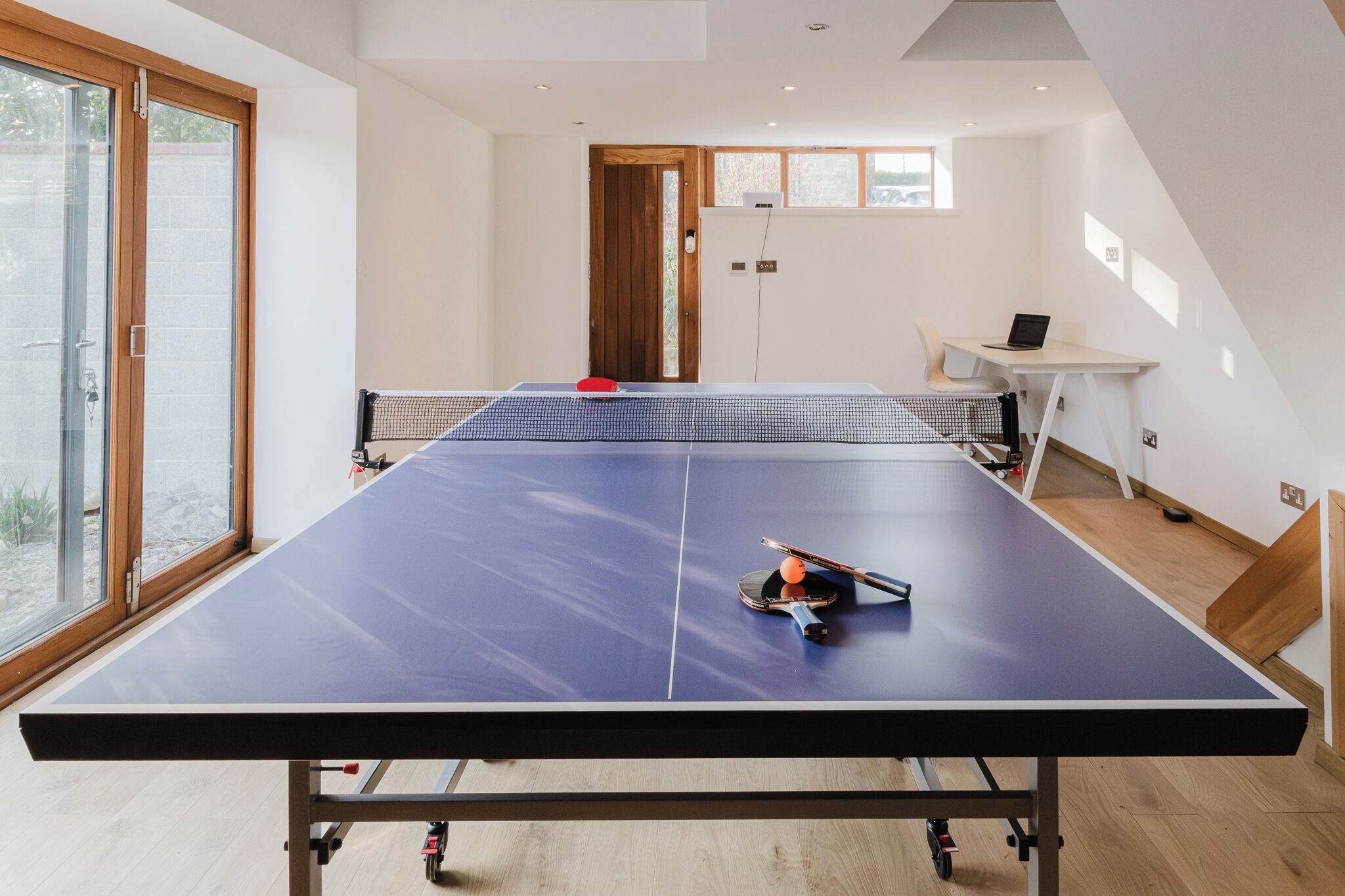Une table de ping-pong se trouve dans une pièce dotée d’une grande fenêtre, d’une porte et d’un bureau avec un ordinateur portable.