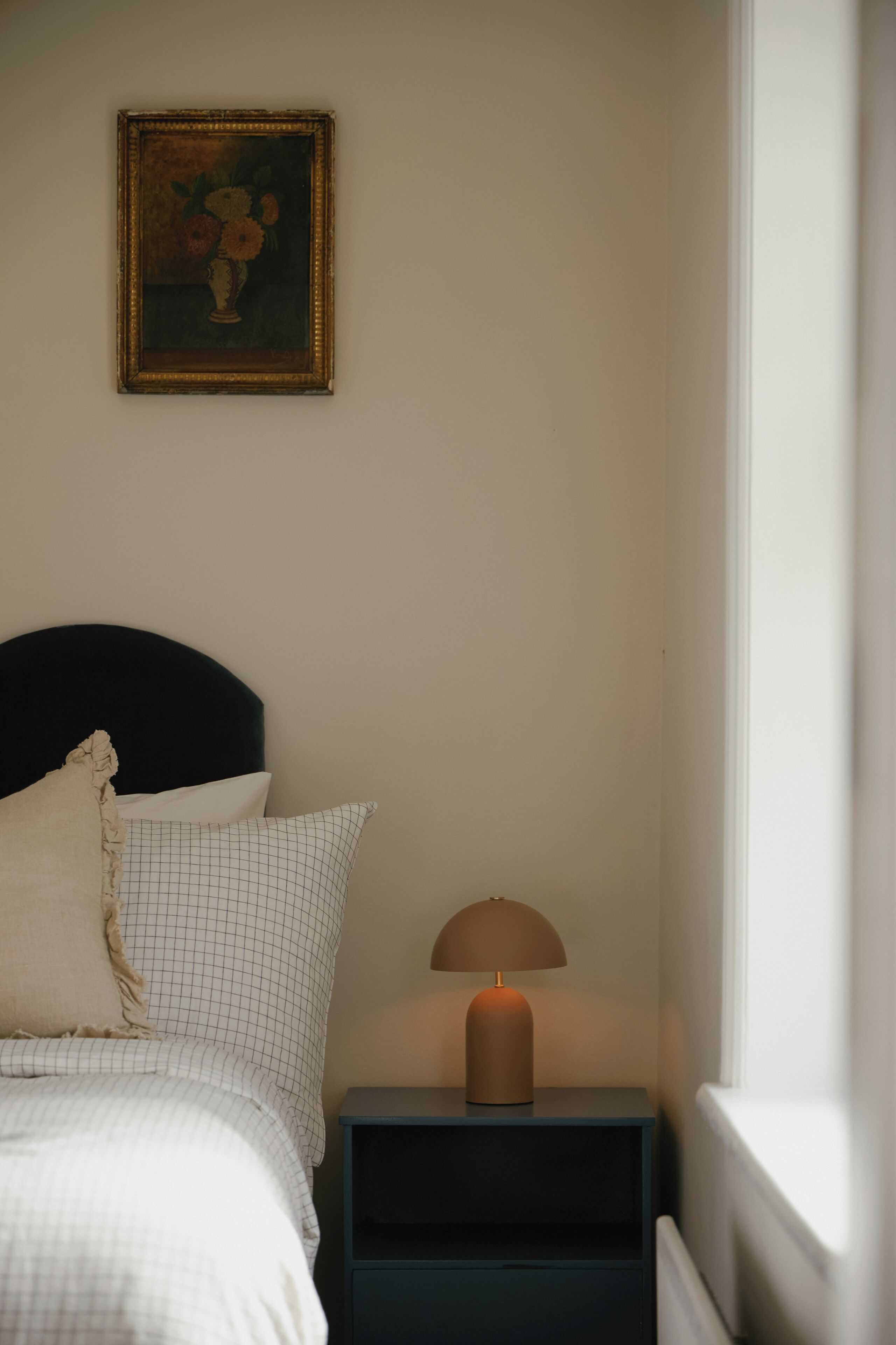 A bedroom with a bed, a nightstand with a lamp, a framed picture on the wall, and a window.