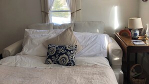 Ein Bett mit weißer Bettwäsche und einem Zierkissen, ein Nachttisch mit einer Lampe, einem gerahmten Bild und einer Wanduhr.