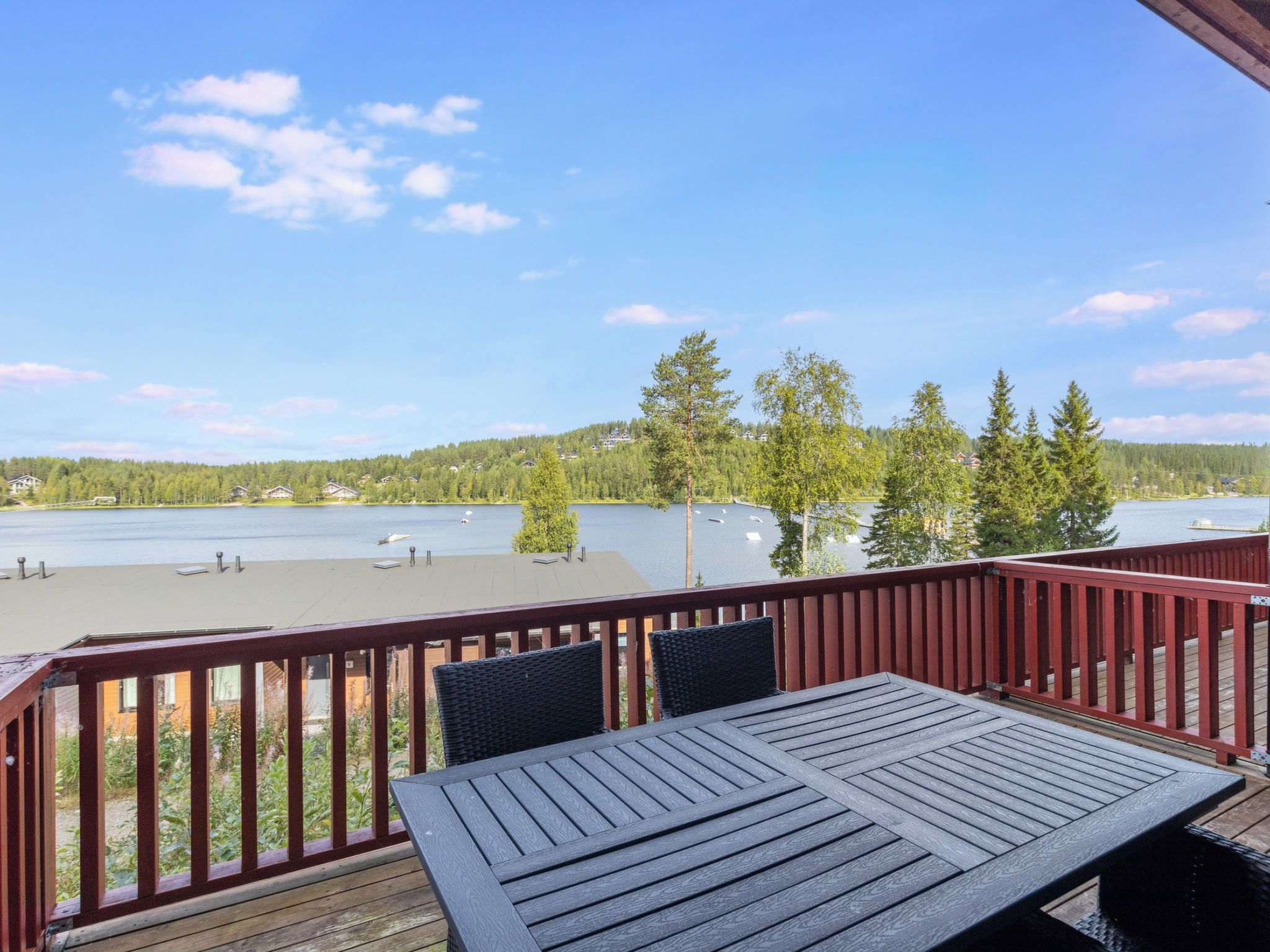 A wooden deck with a table and chairs, overlooking a lake with houses and trees in the background.