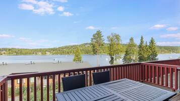 A wooden deck with a table and chairs, overlooking a lake with houses and trees in the background.