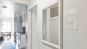 En gang med en hvid dør, der fører ind til et køkken. Der er et indrammet spejl på væggen og et stykke papir med tekst hængende på den.