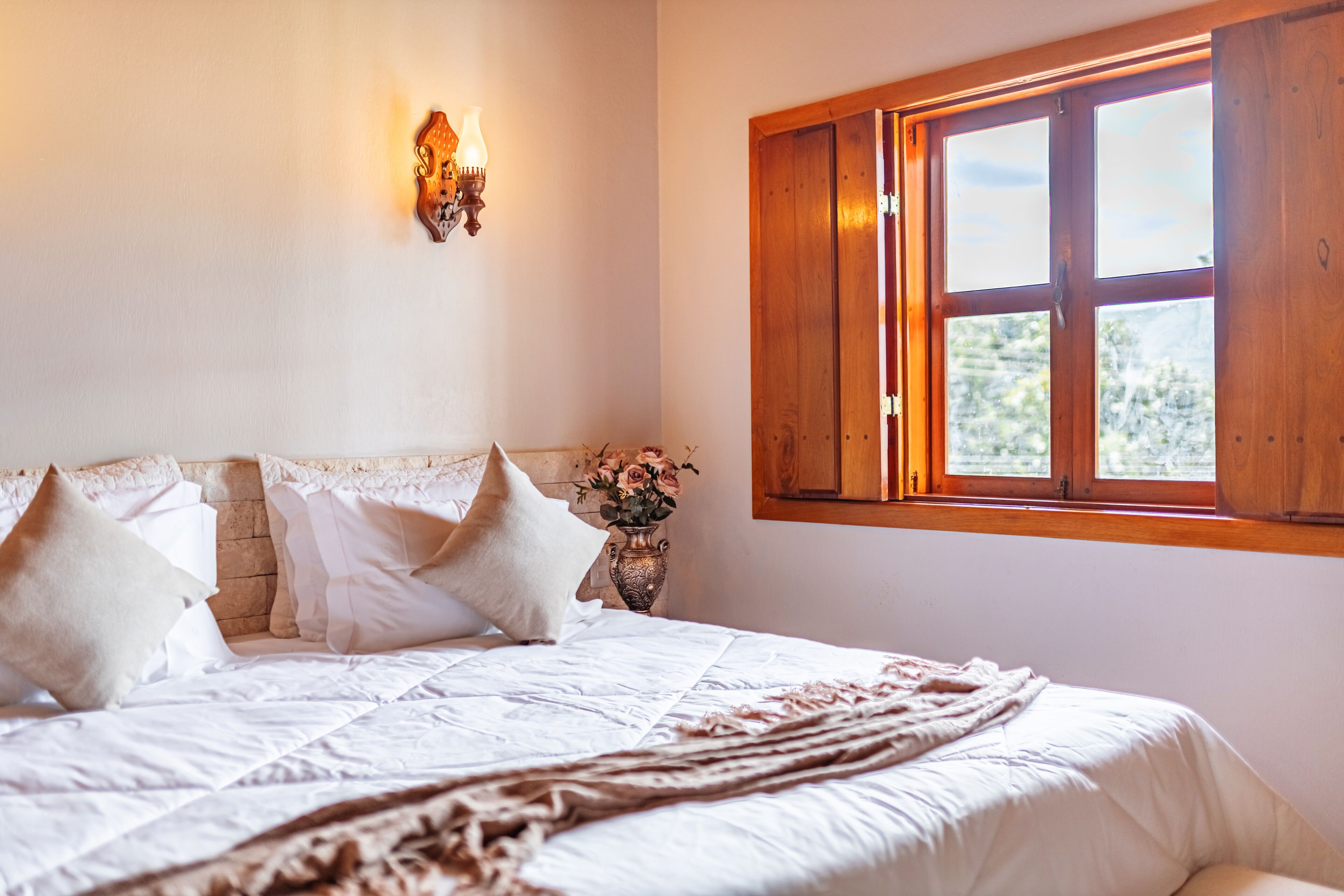 Ein Schlafzimmer mit Bett, Kissen, einer Blumenvase, einer Wandleuchte und einem Fenster mit Holzläden.