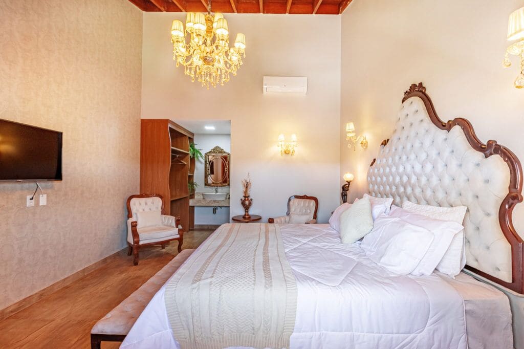 A bedroom with a large bed, a chandelier, a television, and a wooden bench.