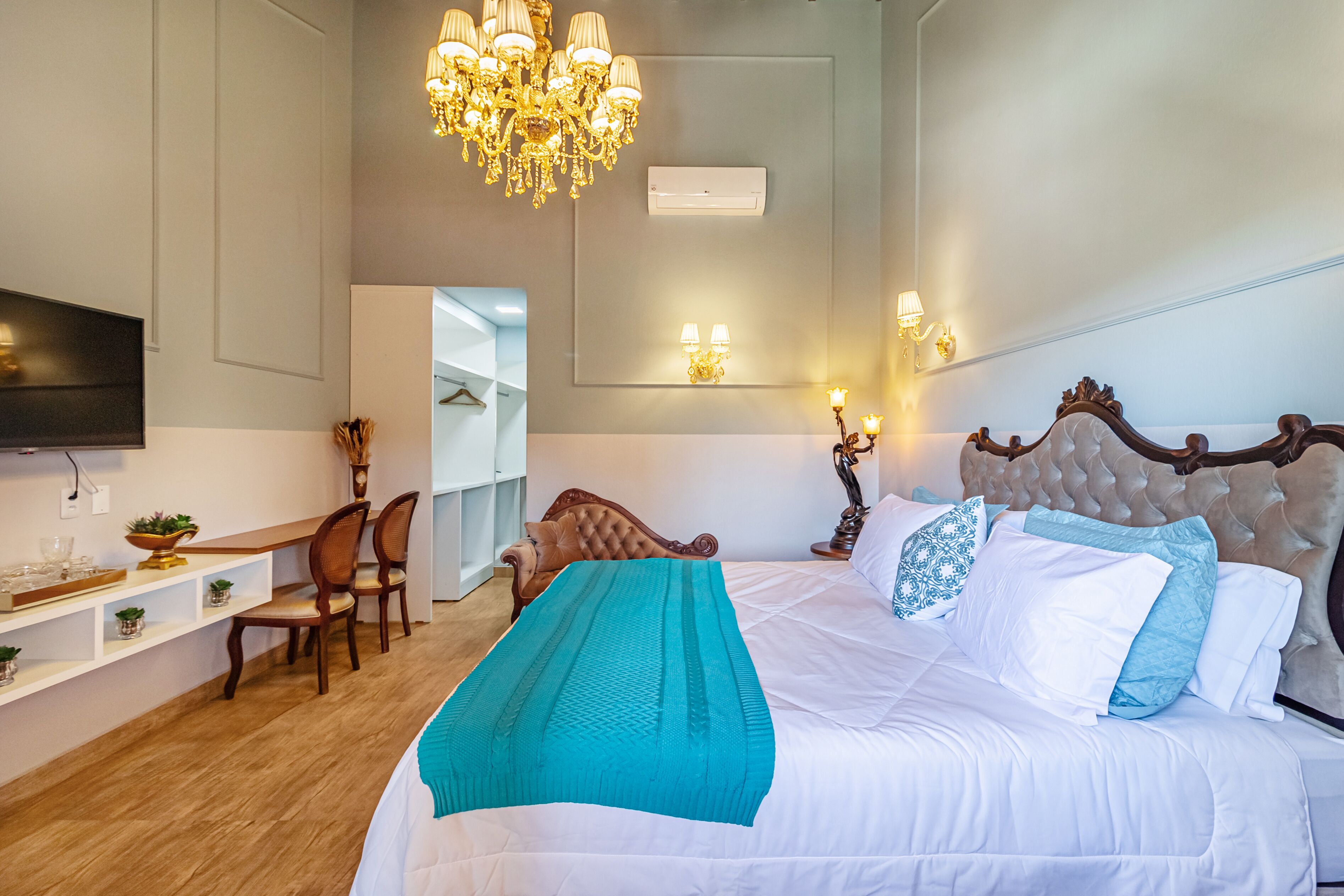 A hotel room with a large bed, a desk with chairs, a television, and a chandelier.