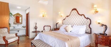 A bedroom with a large bed, a bench, a wooden chair, a vanity with a mirror, and a chandelier.