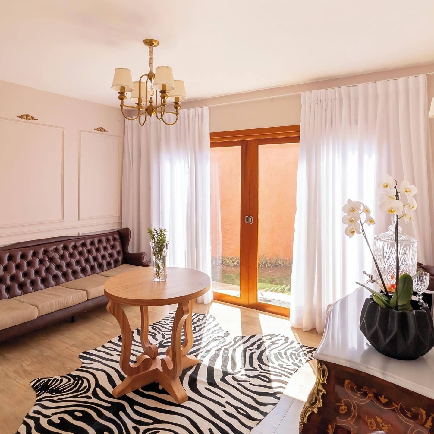 A modern living room with a tufted sofa, a round wooden table, a zebra-patterned rug, and a view through a glass door.